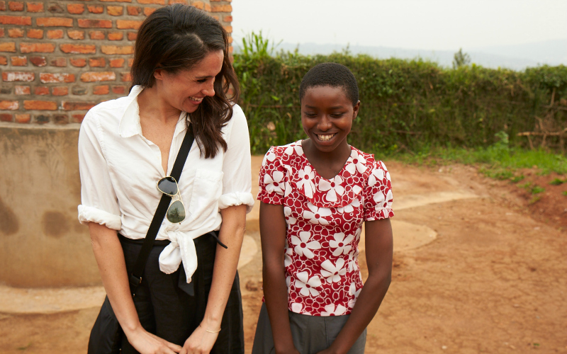 Meghan Markle sits with a child outside in Rwanda, looking across as the child smiles at the camera