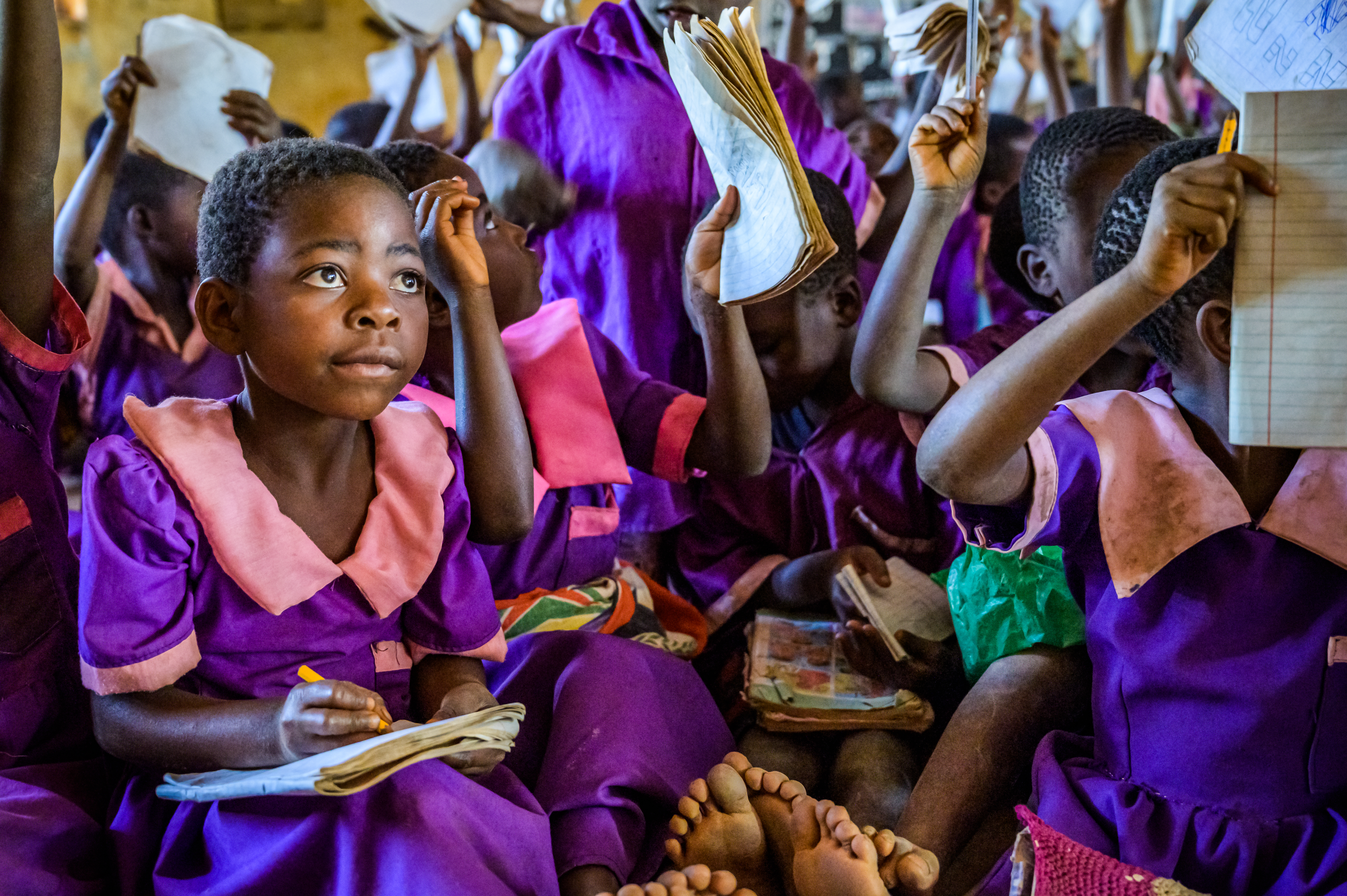 A girl from Malawi in a school uniform with a notebook on her lap listening to the class, with lots of classmates behind her