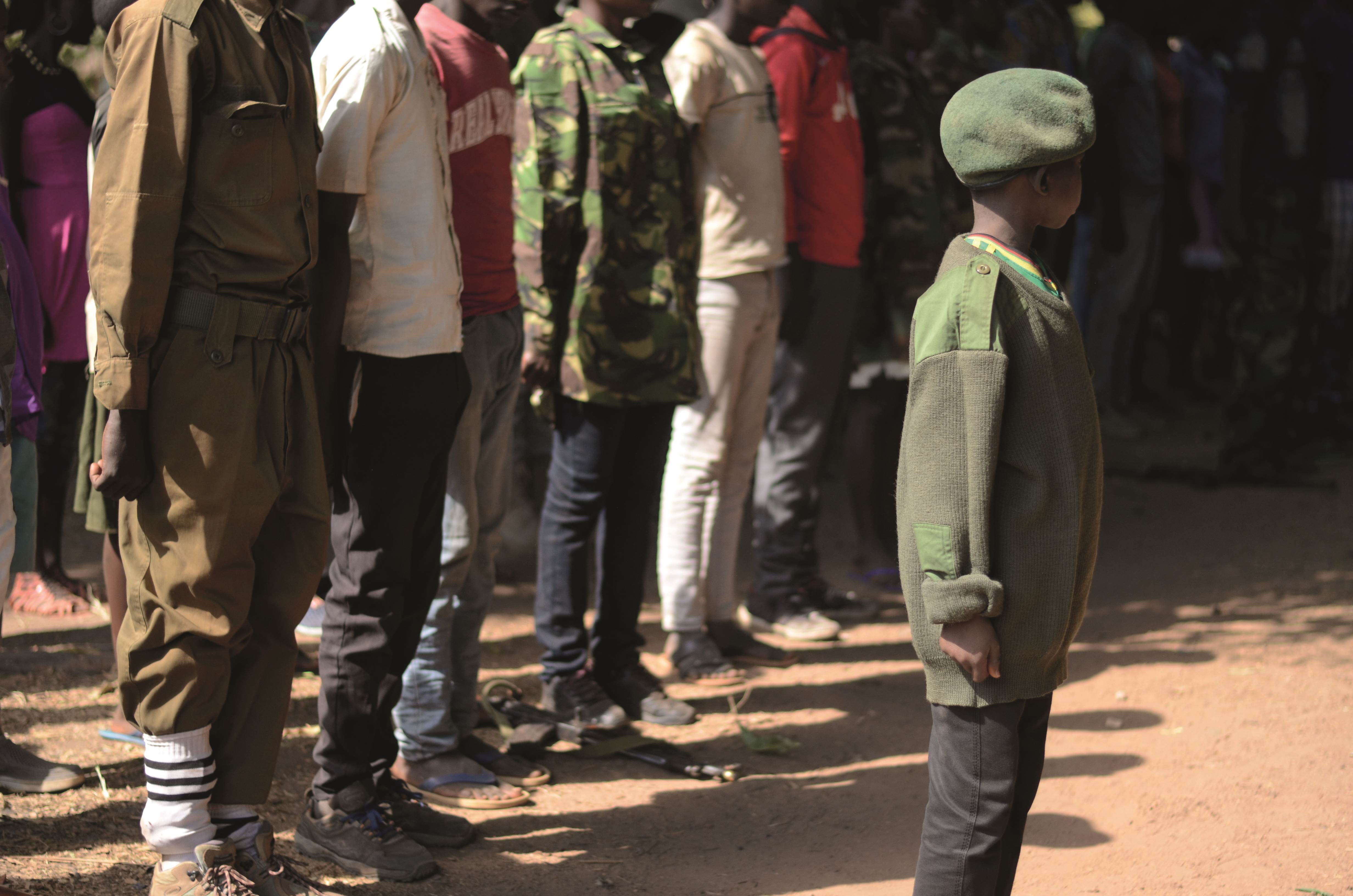 People stand in line behind a child wearing army camouflage and a beret in South Sudan
