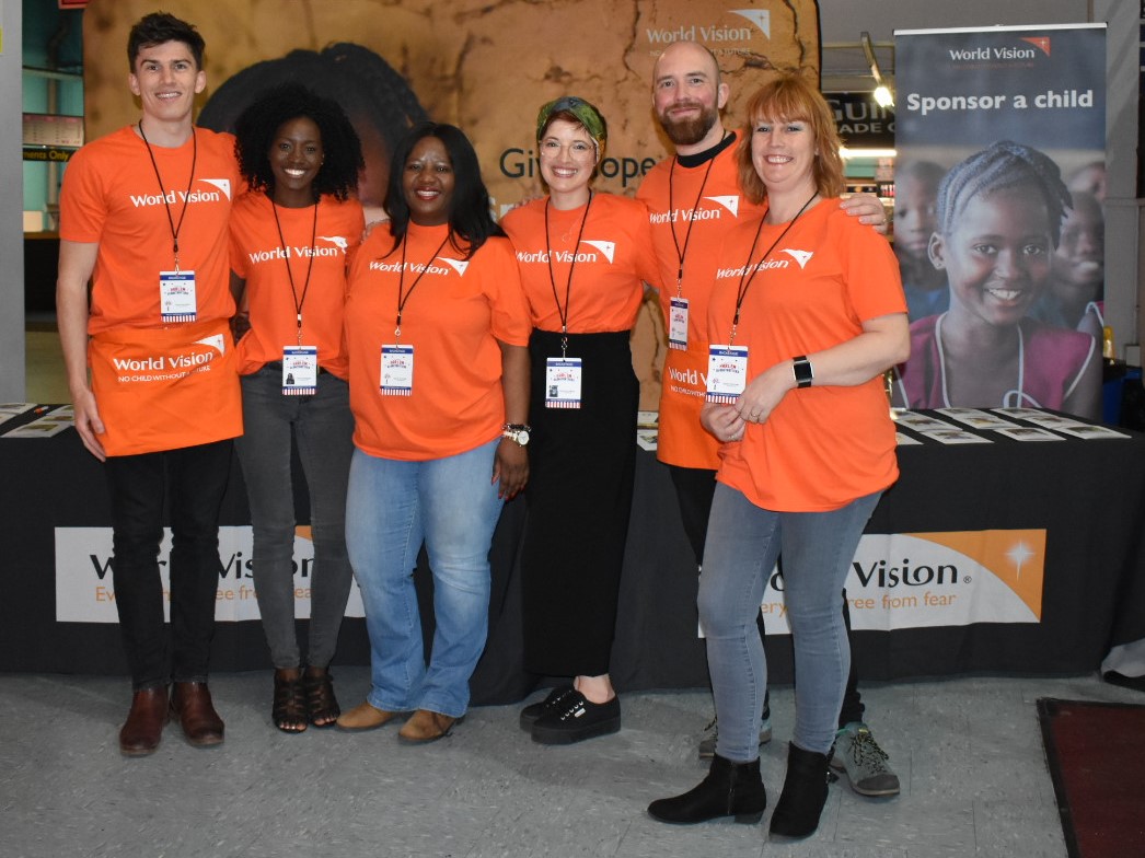 Six World Vision staff in orange tshirts in front of a stand volunteering at a World Vision event