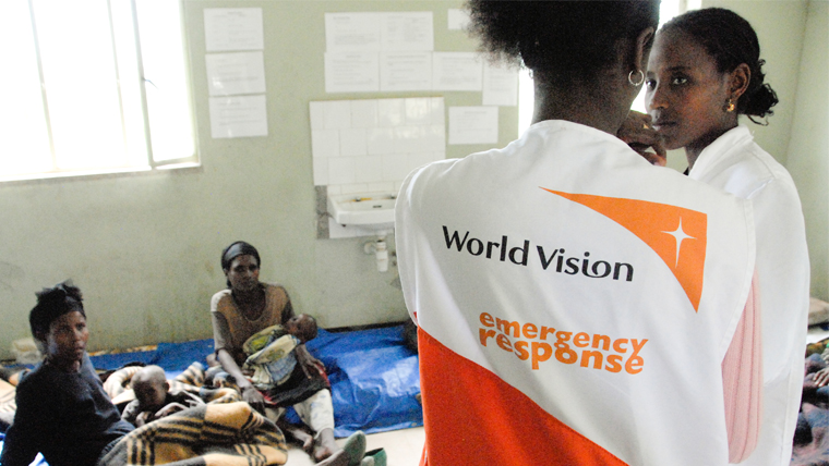 Back of World Vision staff member wearing a tabard that says World Vision Emergency Response in a shelter setting.